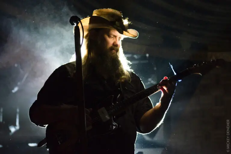 Christ Stapleton plays the guitar with a long beard and cowboy hat at a concert. 