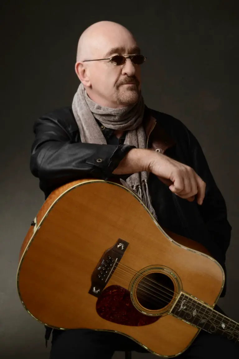 Dave Mason, a bald man with a mustache wearing sunglasses sitting down with an acoustic guitar resting on his leg.