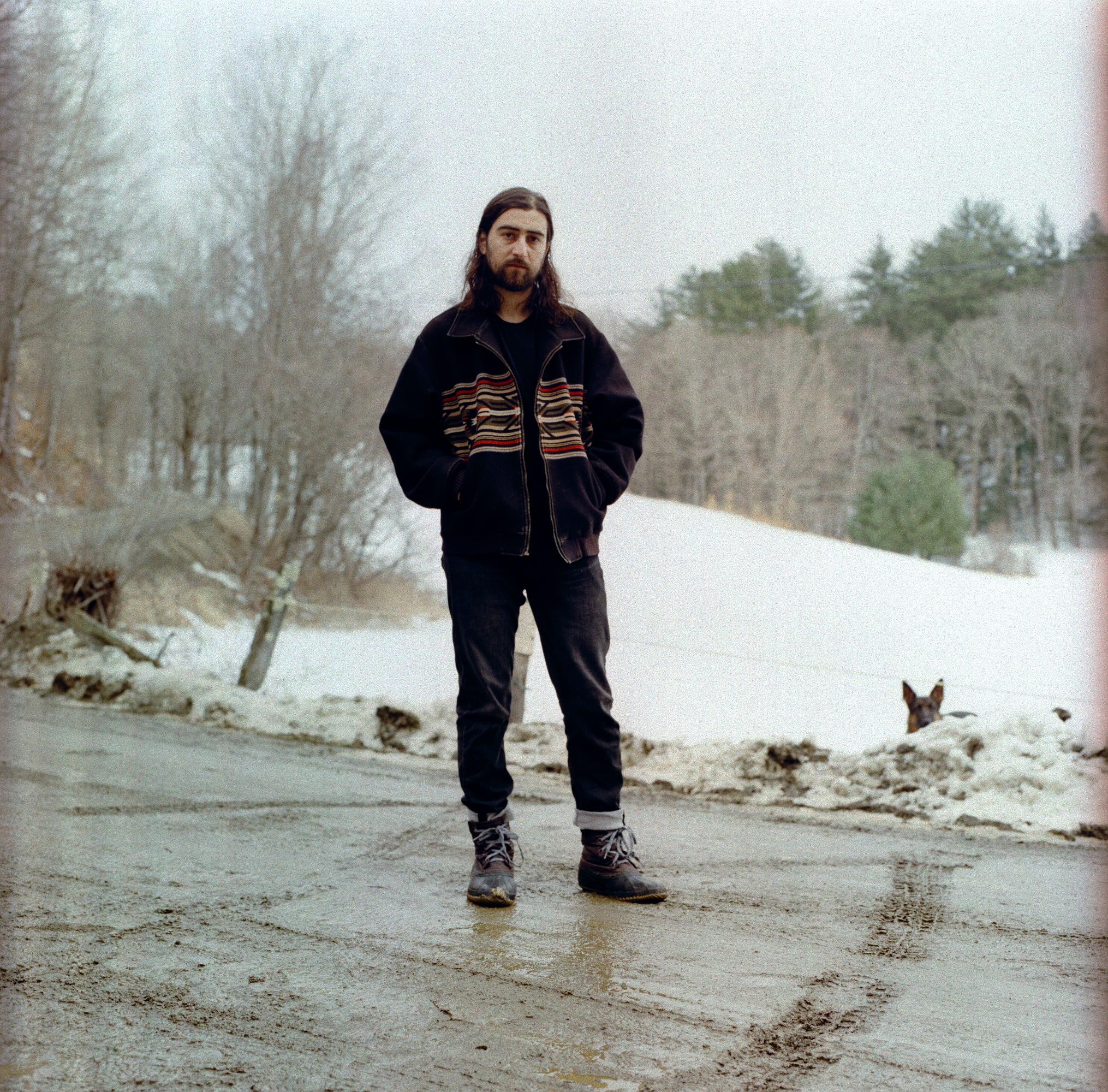 Noah Kahan standing in road with snow behind him and dog peeking head from behind snow pile