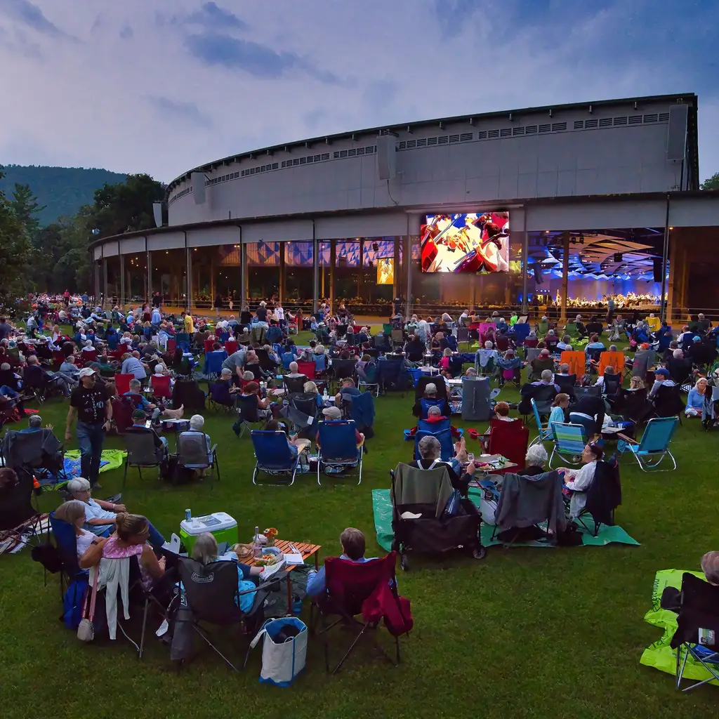 Tanglewood photo from back of the lawn as the sun has set with an orchestra playing on stage and people on lawn chairs in the lawn area